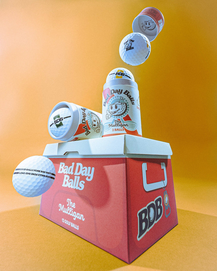 A stylized product render of a red cooler box branded 'Bad Day Balls' with golf balls and cans floating around it against a warm orange gradient background.