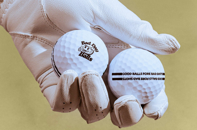 A white leather golf glove holding two white golf balls against a solid olive-green background. One ball features the 'Bad Day Balls' logo, and the other displays custom black printed text.