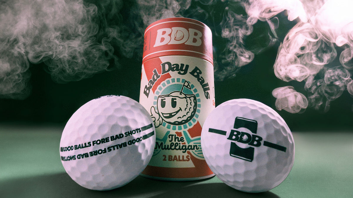 Three BDB branded golf balls and a matching can of 'Bad Day Balls' are arranged on a dark green surface with dramatic smoke effects in the background. One golf ball reads 'BLOOD BALLS FORE BAD SHOTS', another has the BDB logo, and the center can shows the product branding.